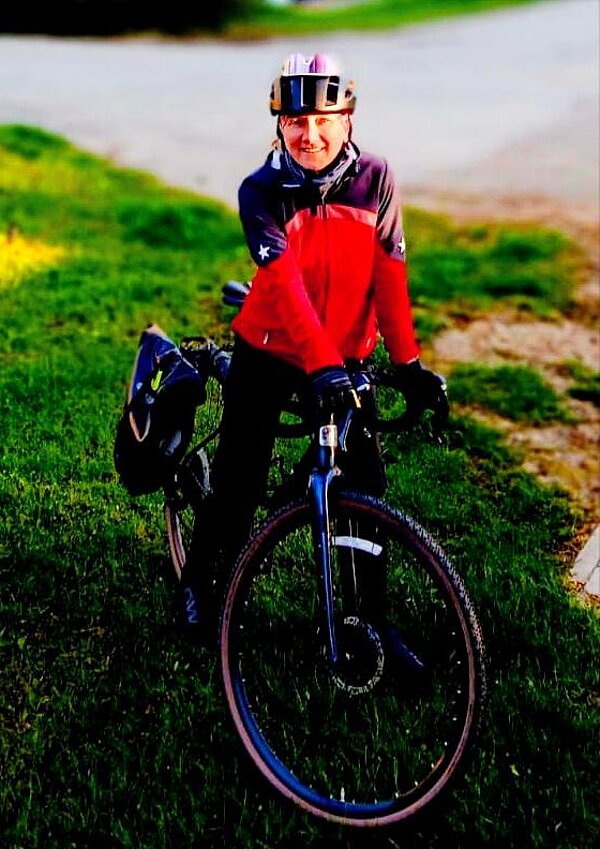 Eine Person steht neben einem Fahrrad auf einer grünen Wiese. Sie trägt einen Helm und eine rot-schwarze Fahrradjacke. Das Fahrrad hat große Reifen, und die Umgebung ist von einer Straße umgeben.