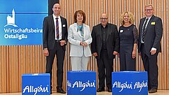 Fünf Personen stehen vor einem Holzpaneel mit dem Schriftzug "Wirtschaftsbeirat Ostallgäu". Sie tragen formelle Kleidung und stehen hinter blauen Kisten mit "Allgäu" darauf. Die Szene vermittelt ein Gefühl von geschäftlicher Zusammenarbeit oder einer Veranstaltung.