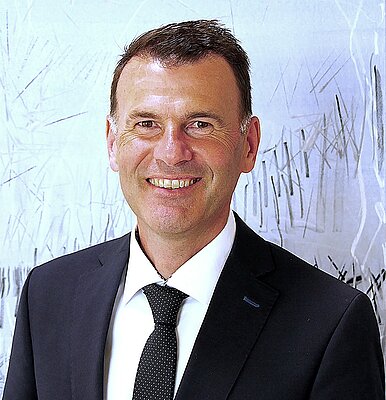 Porträt eines lächelnden Mannes in einem Anzug mit Schemata im Hintergrund. Er hat kurzes, braunes Haar und trägt eine Krawatte. Sein Gesichtsausdruck wirkt freundlich und einladend.