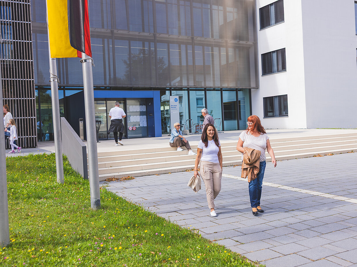 Zwei Frauen gehen lächelnd vor einem modernen Gebäude mit großen Fenstern. Im Hintergrund sind weitere Personen zu sehen, die sich auf Treppen und dem Platz vor dem Gebäude aufhalten. Die Frauen tragen lässige Kleidung und halten Taschen.