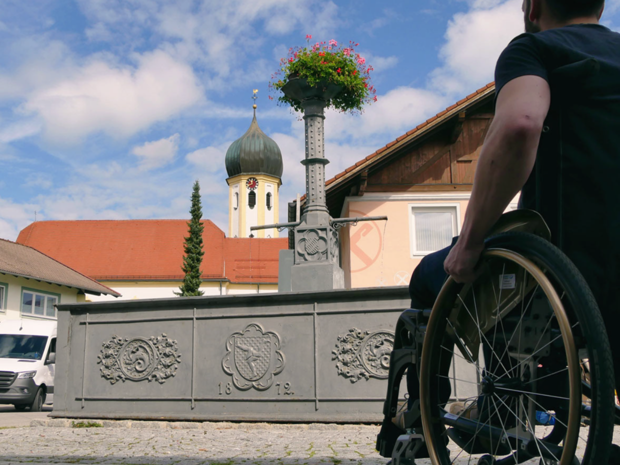 Ein Mann sitzt im Rollstuhl und blickt auf einen Brunnen mit einer Pflanze darauf. Im Hintergrund ist eine Kirche mit einer Kuppel zu sehen. Der Himmel ist blau mit wenigen Wolken.