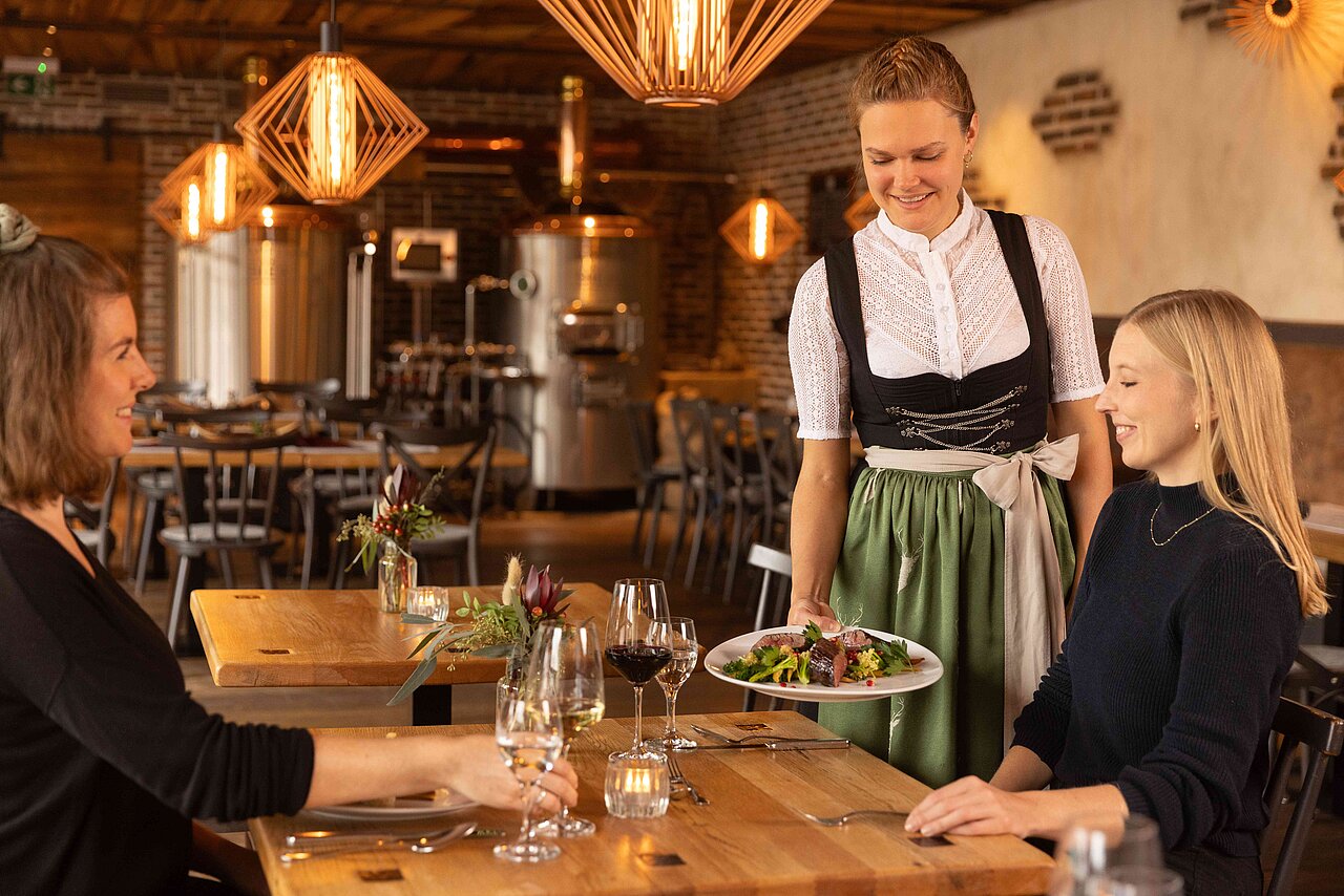 Eine Kellnerin in traditioneller Tracht serviert einem Tisch mit zwei Gästen ein Gericht in einem stilvollen Restaurant. Die Atmosphäre ist gemütlich, mit warmem Licht und modernen Holz-Elementen.