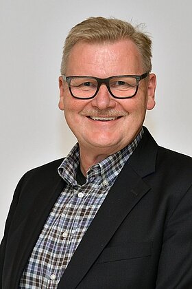 Ein lächelnder Mann mit Brille und einem Hemd im karierten Muster trägt einen blazer. Der Hintergrund ist einfarbig und neutral.