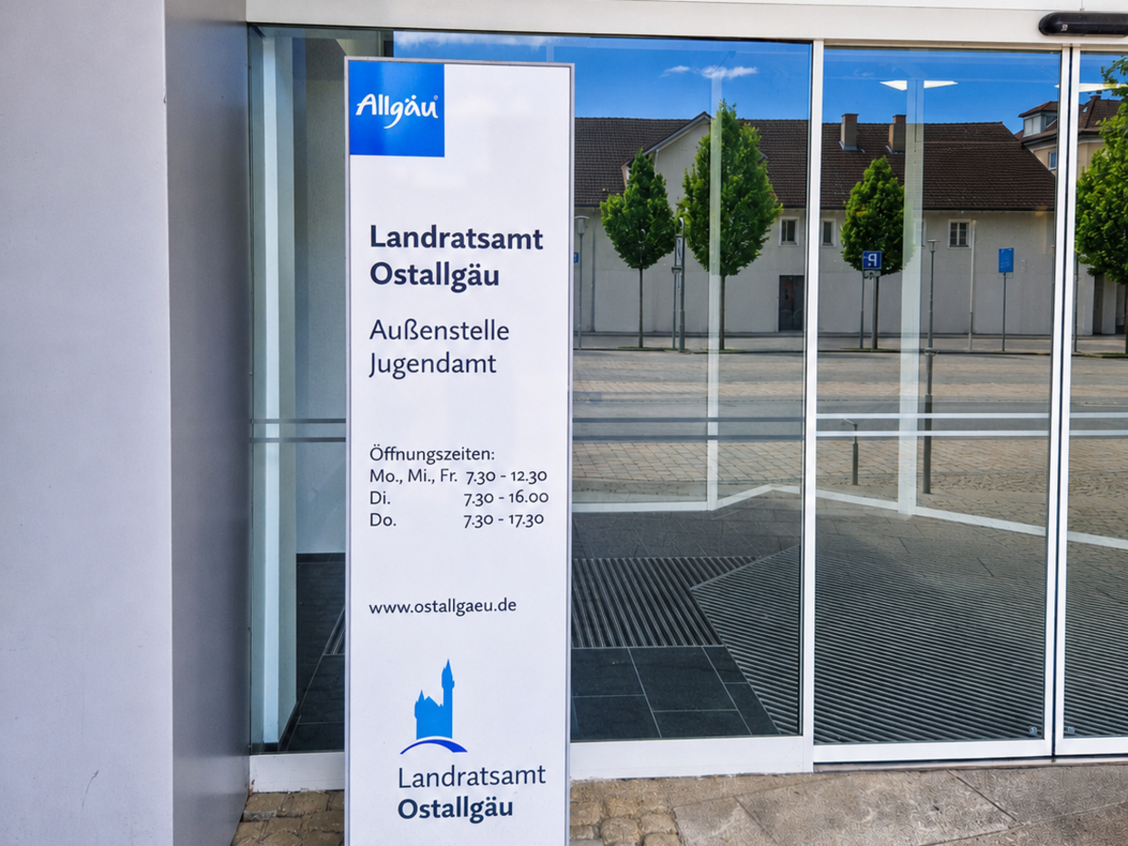 Ein Schild vor einem Gebäude, das das Landratsamt Ostallgäu und die Außenstelle Jugendamt anzeigt. Es enthält Öffnungszeiten: Montag bis Mittwoch von 7:30 bis 12:30, sowie Dienstag und Donnerstag von 7:30 bis 17:30. Das Schild hat ein einfaches, modernes Design.