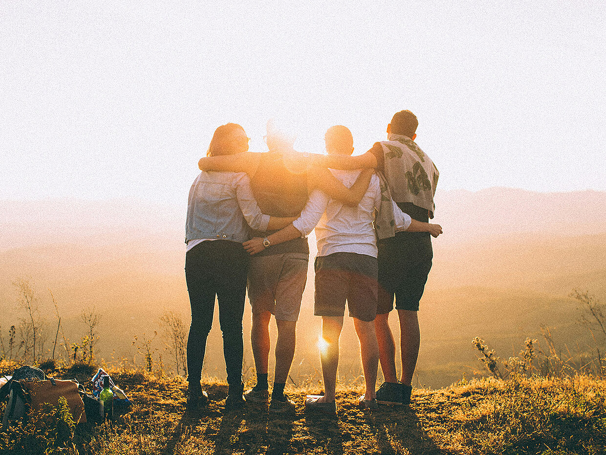 Eine Gruppe von vier Personen steht Rücken an Rücken auf einem Hügel, während die Sonne untergeht. Sie umarmen sich und blicken auf die Landschaft. Die Atmosphäre ist warm und freundschaftlich, mit sanften Lichtstrahlen, die die Szene dornen.