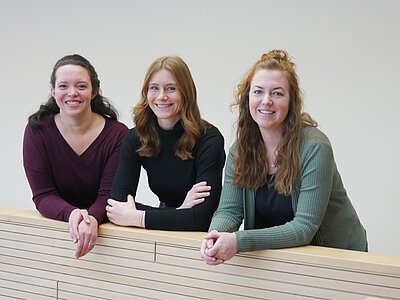 Drei Frauen stehen an einer Stütze und lächeln in die Kamera. Sie tragen unterschiedliche Oberteile in verschiedenen Farben und haben unterschiedliche Frisuren. Die Hintergrundwand ist hell und unauffällig.