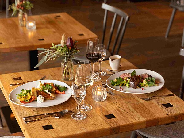 Ein eleganter Tisch in einem Restaurant, gedeckt mit zwei Tellern köstlicher Speisen. Es gibt Salate, Fleischgerichte und dekorative Gläser mit Wein. Kleine Kerzen und Blumenvasen sorgen für eine gemütliche Atmosphäre. Der Holzfußboden und die Einrichtung verleihen dem Raum einen modernen, einladenden Charakter.
