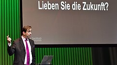 Ein Mann in einem Anzug steht auf einer Bühne und spricht. Im Hintergrund ist eine Präsentation mit der Frage "Lieben Sie die Zukunft?" zu sehen. Der Raum hat grüne Hintergrundbeleuchtung.