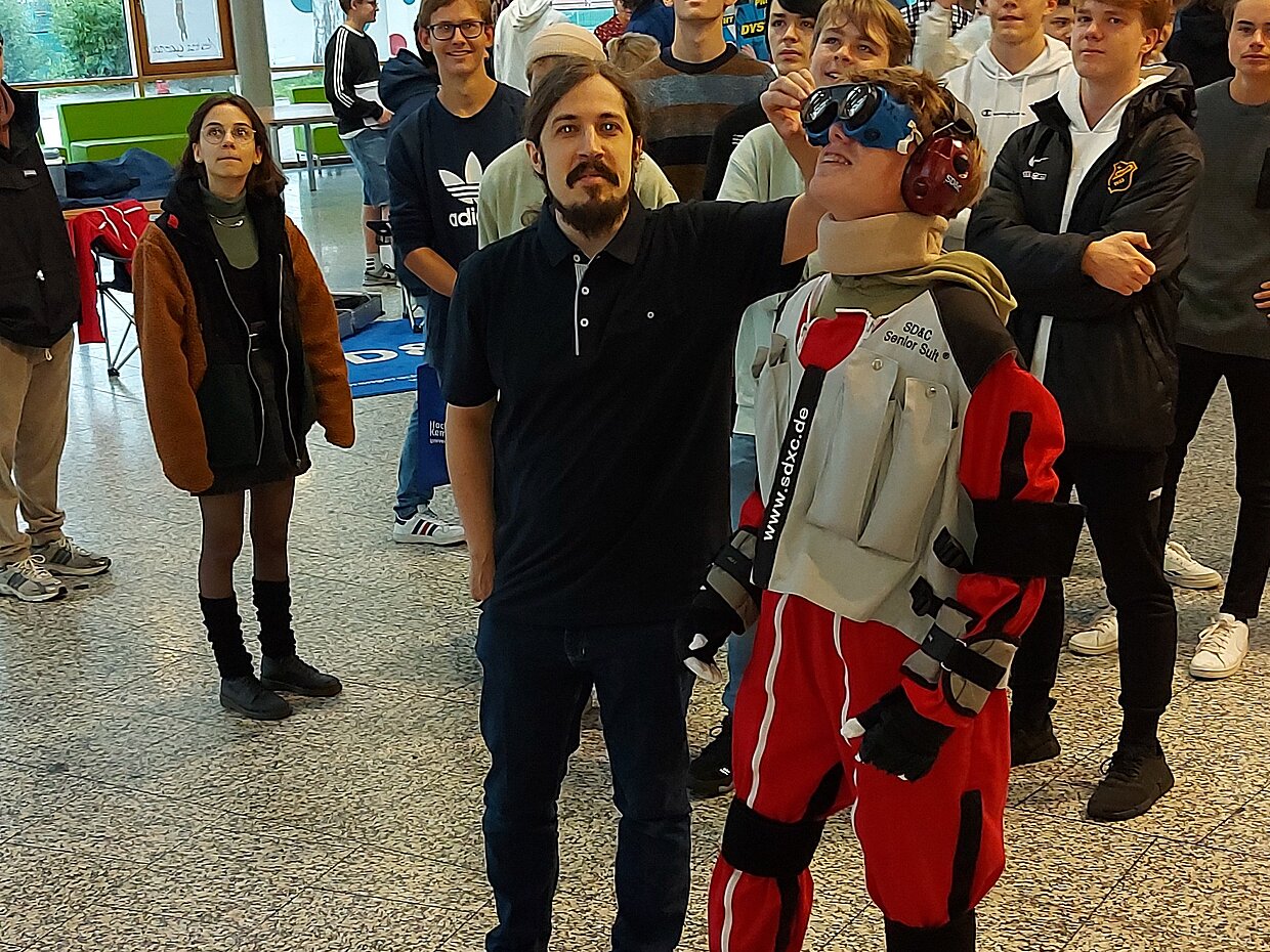 Eine Gruppe von Personen steht in einem Raum. Ein Mann trägt ein schwarzes Shirt und steht neben einer Person in einem auffälligen roten Anzug mit Headset. Im Hintergrund sind weitere Personen zu sehen, die auf etwas schauen.