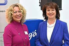 Zwei Frauen stehen lächelnd nebeneinander. Die Frau links trägt ein pinkes Oberteil, die Frau rechts ein blaues Blazerensemble. Im Hintergrund ist ein blauer Gegenstand und eine Banner mit Aufschrift zu sehen. Beide Frauen scheinen an einem Event teilzunehmen.