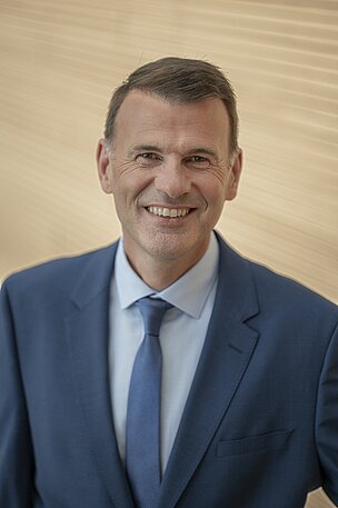 Ein Mann mit kurzem, braunem Haar trägt einen blauen Anzug und ein hellblaues Hemd mit einer gestreiften Krawatte. Er lächelt freundlich und steht vor einem hellen, unscharfen Hintergrund.