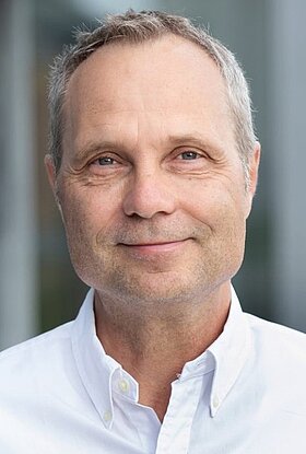 Ein lächelnder Mann mit grauen Haaren trägt ein weißes Hemd. Der Hintergrund ist unscharf und zeigt diffuse, helle Farben. Die Stimmung des Bildes ist freundlich und einladend.