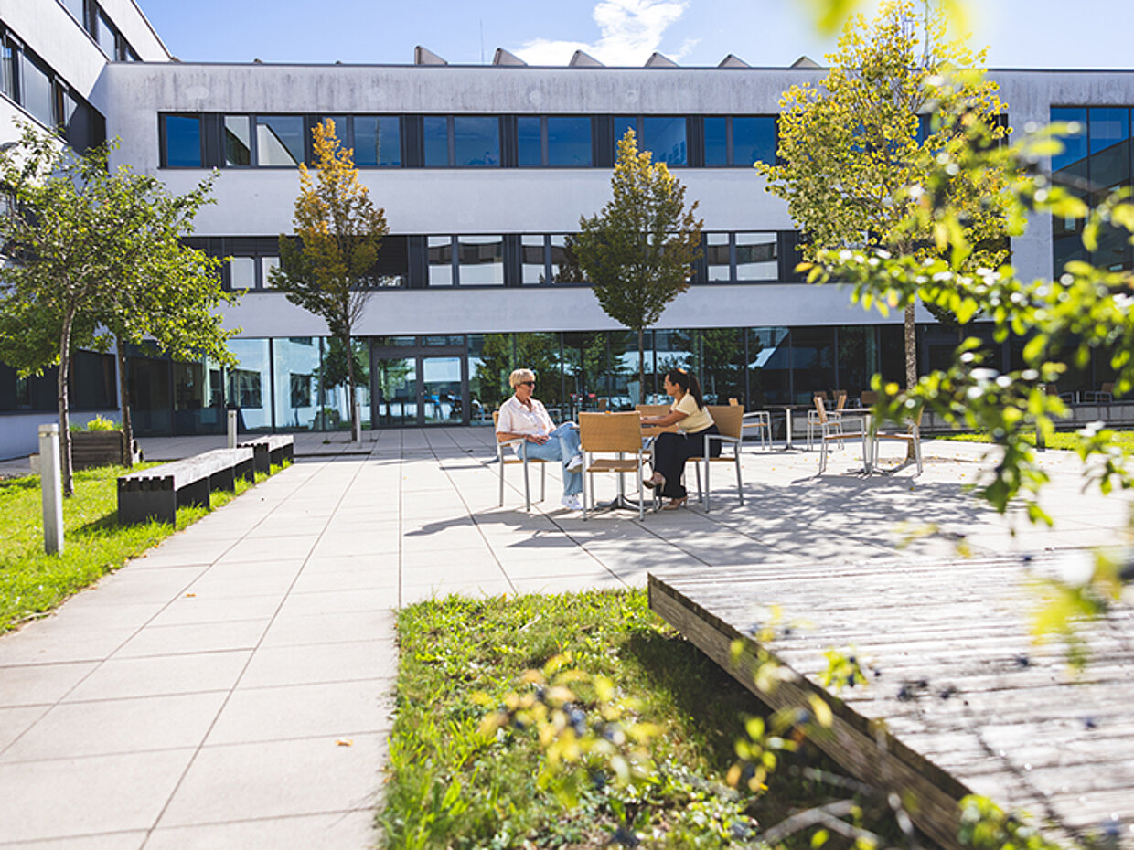 Zwei Personen sitzen an einem Tisch in einem Außenbereich, umgeben von Pflanzen und Bäumen. Im Hintergrund sind Fenster eines modernen Gebäudes sichtbar, und der Himmel ist klar. Der Platz ist mit hellen Fliesen gestaltet.