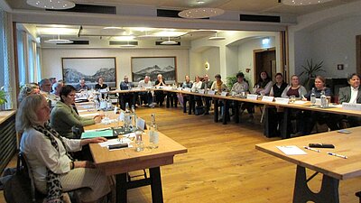 Eine Gruppe von Menschen sitzt an einem langen Tisch in einem Besprechungsraum. Einige haben Notizen, Wasserflaschen stehen bereit. Im Hintergrund befinden sich zwei große Bilder von Berglandschaften. Der Raum ist hell erleuchtet.