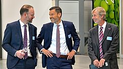 Drei Männer stehen in einem modernen, hellen Raum und unterhalten sich. Sie tragen formelle Anzüge und scheinen in einer freundlichen Stimmung zu sein. Im Hintergrund sind unscharfe, grüne Elemente zu erkennen.
