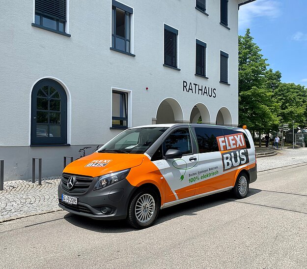 Flexibus Marktoberdorf Ein orange-weißer Kleinbus mit der Aufschrift "FLEXI BUS" steht vor einem Rathausgebäude. Der Bus ist ein Elektrofahrzeug und auf einem gepflasterten Weg geparkt, umgeben von Bäumen.