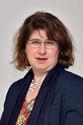 Porträt einer Frau mit brünettem, lockigem Haar, die eine Brille trägt. Sie hat ein freundliches Lächeln und trägt eine modische Bluse mit buntem Muster sowie einen dunklen Blazer und eine Perlenkette. Der Hintergrund ist neutral hell.
