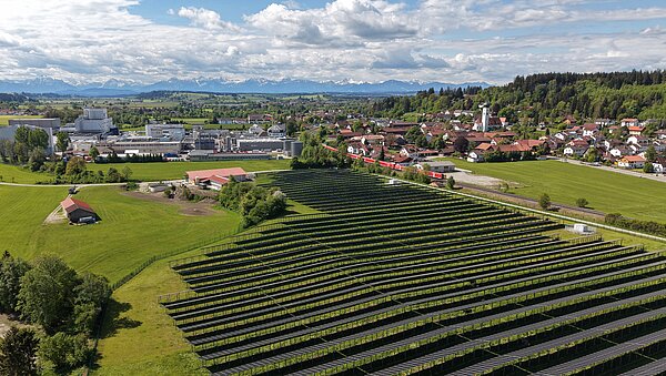 Luftaufnahme einer ländlichen Gegend mit einem großen Feld von Solarmodulen im Vordergrund. Im Hintergrund sind Wohnhäuser, Gebäude und eine Landschaft mit Bergen in der Ferne zu sehen. Das Bild vermittelt einen Eindruck von erneuerbarer Energie und ländlicher Idylle.