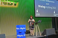 Eine Frau mit rotem Haar steht am Rednerpult auf einer Bühne. Im Hintergrund ist ein Bildschirm, auf dem „Herzlich Willkommen zum Dialogempfang 2022“ steht. Links neben der Frau sind blaue Kisten, auf denen „Allgäu“ geschrieben steht.