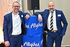 Zwei Männer in Anzügen lächeln und stehen neben blauen Würfeln mit der Aufschrift „Allgäu“. Sie zeigen eine freundliche Haltung und scheinen sich in einem Veranstaltungsraum zu befinden. Im Hintergrund sind unscharfe Veranstaltungselemente zu erkennen.