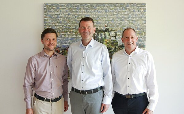Pirmin Joas (Bayer. Gemeindetag, stv. Kreisverbandsvorsitzender), Bernd Stapfner (Landrat), Helmut Bucher (Bayer. Gemeindetag,Kreisverbandsvorsitzender) Drei Männer stehen freundlich nebeneinander vor einem bunten, abstrakten Gemälde. Der Mann links trägt ein helles Hemd, der in der Mitte ein weißes Hemd und der rechts ein ähnliches Hemd. Alle haben eine entspannte Körperhaltung und lächeln.