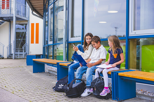 Drei Kinder sitzen auf einer Bank vor einem Schulgebäude. Sie betrachten gemeinsam ein Heft und lachen. Zwei Jungen tragen T-Shirts und Jeans, das Mädchen hat lange Haare und ein lila T-Shirt an. Es sind Schulranzen neben ihnen auf dem Boden.