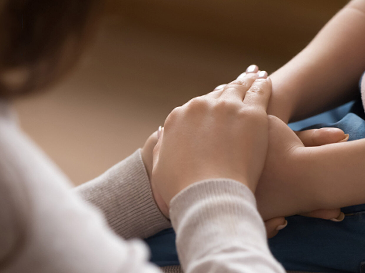 Zwei Hände halten sich sanft. Eine Hand ist auf die andere gelegt, was Nähe und Unterstützung symbolisiert. Die Umgebung ist unscharf, was den Fokus auf die Hände lenkt.