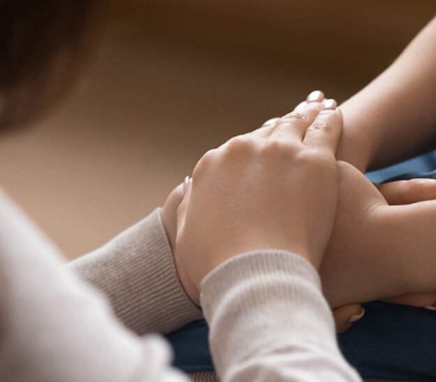 Zwei Hände halten sich sanft. Eine Hand ist auf die andere gelegt, was Nähe und Unterstützung symbolisiert. Die Umgebung ist unscharf, was den Fokus auf die Hände lenkt.