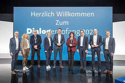 Eine Gruppe von neun Personen steht auf einer Bühne vor einem großen, blauen Banner mit der Aufschrift "Herzlich Willkommen Zum Dialogempfang 2022" und dem Logo von DMG MORI. Die Personen treten formal auf und lächeln in die Kamera.
