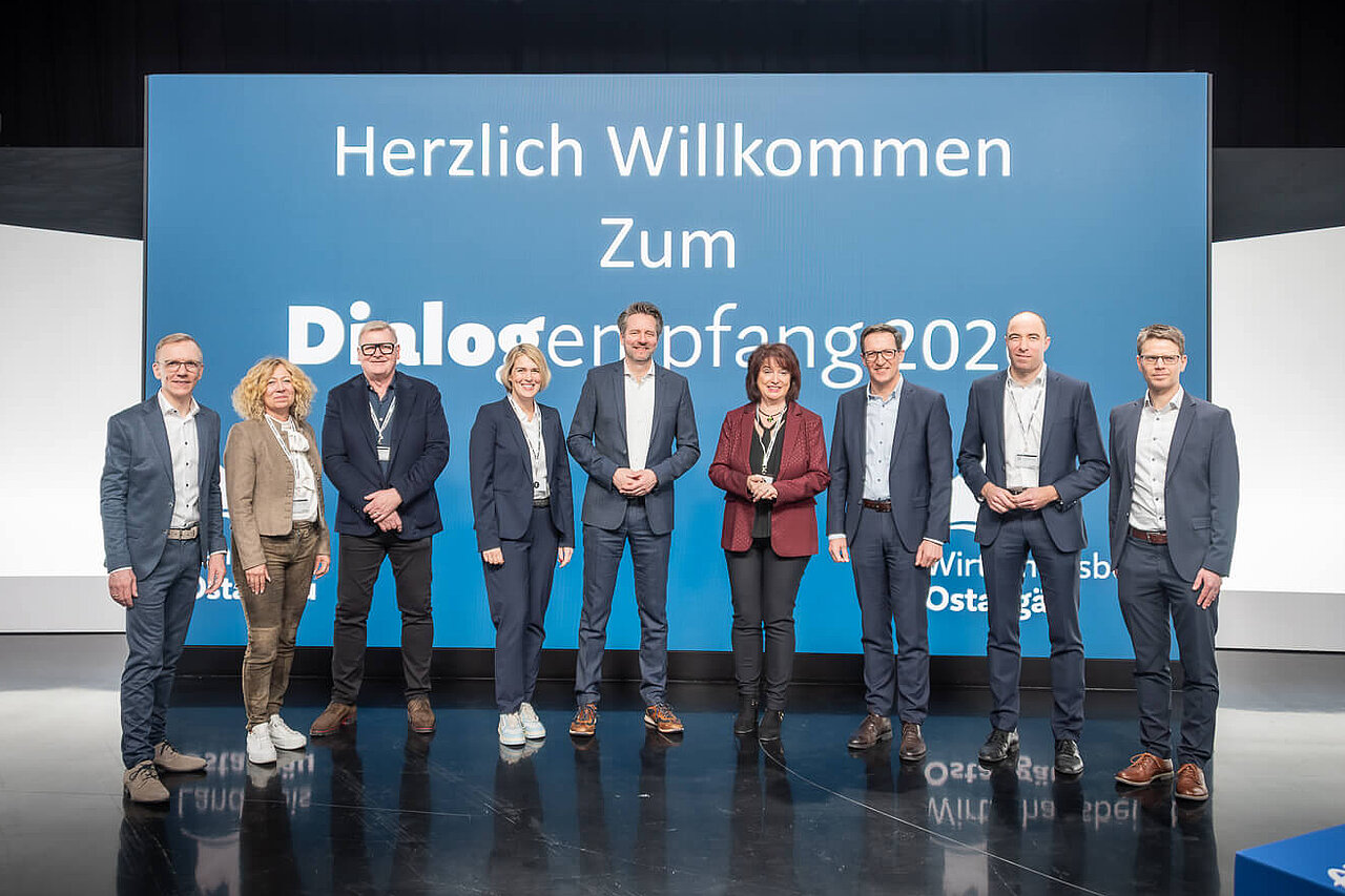 Eine Gruppe von neun Personen steht auf einer Bühne vor einem großen, blauen Banner mit der Aufschrift "Herzlich Willkommen Zum Dialogempfang 2022" und dem Logo von DMG MORI. Die Personen treten formal auf und lächeln in die Kamera.