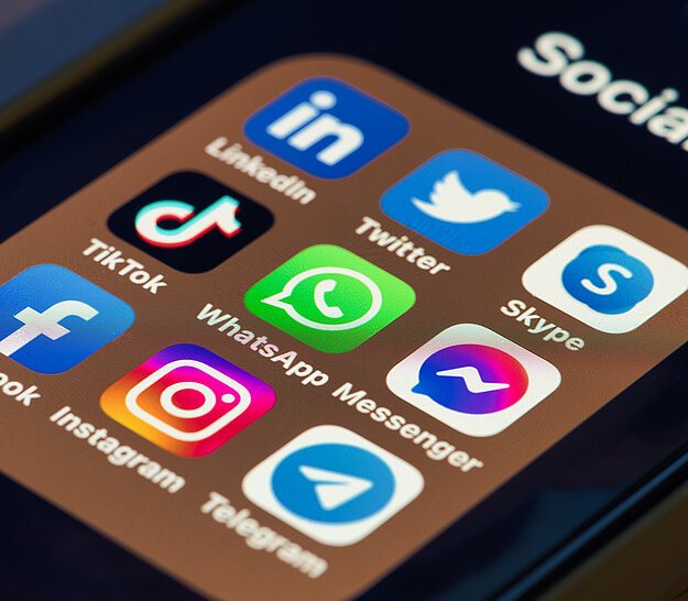 Ein Smartphone-Bildschirm zeigt eine Sammlung von sozialen Medien-App-Icons in einem Ordner mit der Bezeichnung "Social". Zu sehen sind unter anderem Instagram, Facebook, WhatsApp, TikTok, Messenger und Telegram. Der Bildschirm ist unscharf, der Hintergrund zeigt eine Tastatur.