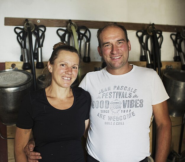 Ein Mann und eine Frau stehen nebeneinander und lächeln in die Kamera. Im Hintergrund sind mehrere Melkgeräte aufgehängt. Die Frau trägt ein schwarzes T-Shirt, der Mann ein weißes T-Shirt mit Aufdruck.
