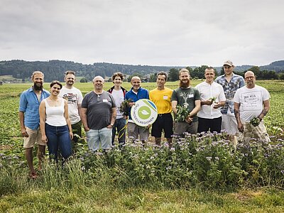 Eine Gruppe von zwölf Menschen steht auf einem Feld, umgeben von Pflanzen. Sie lächeln und halten einige frische Kräuter in der Hand. In der Mitte ist ein großes Schild mit einem grünen Logo zu sehen. Der Himmel ist bewölkt und die Landschaft erstreckt sich im Hintergrund.