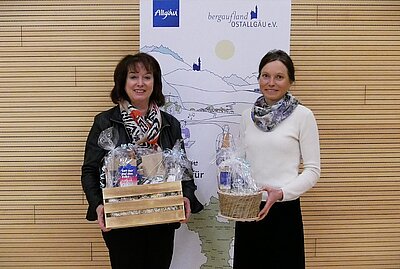 Zwei Frauen stehen vor einer Holzwand und halten Geschenkkörbe in den Händen. Die Frau links trägt eine schwarze Jacke, die Frau rechts ein weißes Oberteil und einen Schal. Im Hintergrund ist eine Karte mit dem Schriftzug "Allgäu" und "Ostallgäu e.V." zu sehen.