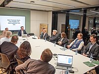 Eine Gruppe von Fachleuten sitzt um einen großen Tisch in einem modernen Besprechungsraum. An der Wand ist eine Präsentation mit dem Titel "Best Practices Smart-City Projekte und Übersichtlichkeit auf QA" zu sehen. Wasserflaschen und Laptops sind auf dem Tisch verteilt.