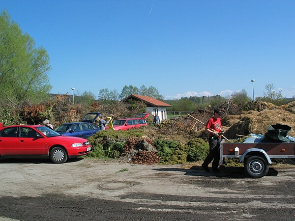 Eine Person steht neben einem Anhänger auf einem Schotterplatz, umgeben von mehreren Autos und einem großen Haufen aus Gartenabfällen. Im Hintergrund sind weitere Personen und eine Holzhütte zu sehen. Der Himmel ist blau und es ist sonnig.