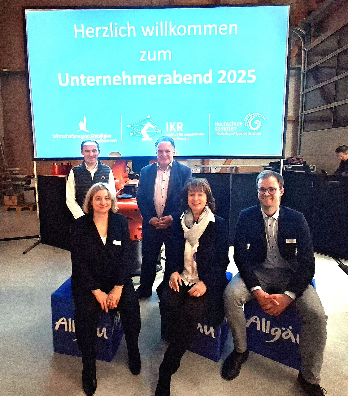 Gruppe von fünf Personen, die vor einer großen Leinwand stehen, auf der „Herzlich willkommen zum Unternehmerabend 2025“ geschrieben steht. Die Umgebung ist eine Veranstaltungshalle mit technischem Equipment. Einige Personen tragen Namensschilder.