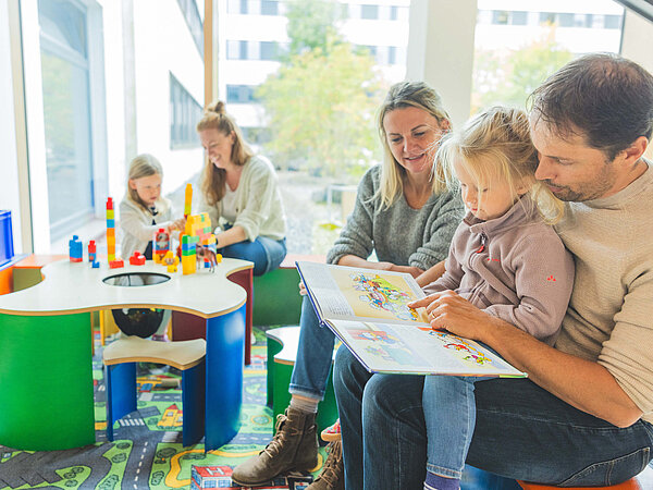 Eine Familie sitzt in einem hellen Raum mit großen Fenstern. Ein Vater liest einem kleinen Mädchen in einem Buch vor, während die Mutter daneben sitzt. Im Hintergrund spielen zwei weitere Kinder am Tisch mit buntem Spielzeug. Boden mit Teppich, der eine Straßenkarte zeigt.