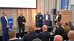 Eine Präsentation auf einer Veranstaltung mit mehreren Sprechern auf der Bühne. Im Vordergrund sitzen Zuhörer, die dem Vortrag aufmerksam folgen. Auf der Bühne steht ein Redner am Mikrofon, während andere sich auf Notizen konzentrieren.