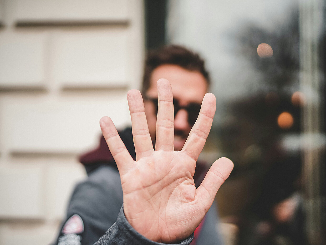Eine Person steht im Vordergrund und hält ihre Hand mit den Fingern nach vorne, um eine Geste der Ablehnung oder des Stoppens auszudrücken. Im Hintergrund sind unscharfe Lichter und eine Wand zu sehen.
