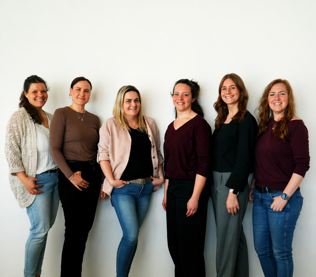 Sechs Frauen stehen in einer Reihe vor einer hellen Wand. Jede Frau trägt unterschiedliche Kleidungsstile, darunter Pullis und Jeans. Die Atmosphäre wirkt freundlich und professionell, mit Lächeln auf den Gesichtern der Frauen.