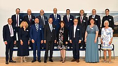 Gruppenfoto von 17 Personen in formeller Kleidung, die in zwei Reihen stehen und sitzen. Hintergrund zeigt eine landschaftliche Fotografie. Jeder trägt ein Namensschild, einige haben verschiedene Looks, darunter Anzüge und elegante Kleider.