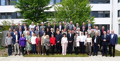 Gruppenfoto von etwa 50 Personen im Freien, vor einem modernen Gebäude und einer größeren Baumlandschaft. Die Teilnehmenden tragen überwiegend formelle Kleidung und stehen in verschiedenen Posen, einige lächeln in die Kamera.