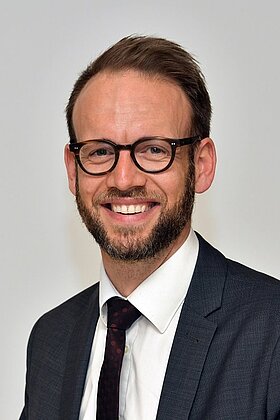 Ein Mann mit Brille und Bart lächelt freundlich in die Kamera. Er trägt einen dunklen Anzug mit einer weißen Hemd und einer Krawatte. Der Hintergrund ist neutral und unauffällig.