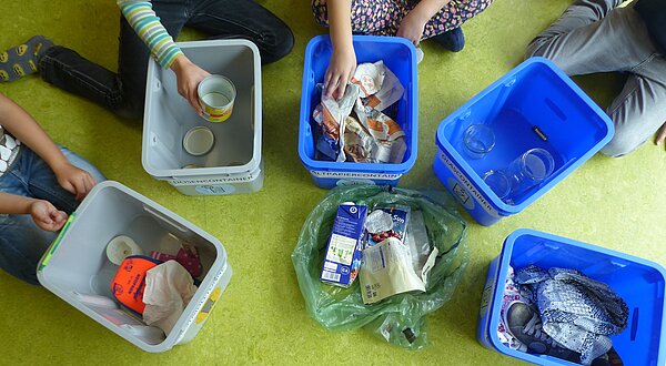 Mehrere Kinder sitzen auf dem Boden und sortieren Müll in verschiedene blaue Behälter. Ein Behälter ist mit leeren Dosen gefüllt, während andere Papier- und Textilabfälle enthalten. Ein grüner Sack liegt in der Mitte. Der Boden ist grün, was auf eine Umgebung in einem Klassenraum hindeutet.