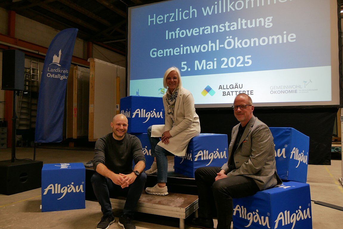 Drei Personen sitzen auf einer Holzplattform vor einem großen Bildschirm, der mit „Herzlich willkommen! Informationsveranstaltung Gemeinwohl-Ökonomie 5. Mai 2025“ beschriftet ist. Um sie herum stehen blaue Boxen mit dem Aufdruck „Allgäu“. Im Hintergrund sind banners und eine einfache Veranstaltungsumgebung sichtbar.