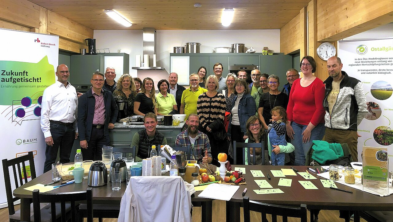 Eine Gruppe von etwa 30 Personen posiert in einer Küche, umgeben von Tischen mit Getränken und Obst. Im Hintergrund stehen Schränke und Kochutensilien. Links und rechts befinden sich Informationsbanner. Alle Mitglieder scheinen an einer Veranstaltung teilzunehmen.