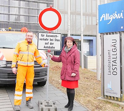 Ein Mann in orangefarbener Arbeitskleidung und eine Frau in rotem Mantel stehen neben einem Verkehrszeichen mit einem Verbotsschild. Im Hintergrund sind ein Fahrzeug und ein Schild des Landratsamtes Ostallgäu sichtbar.