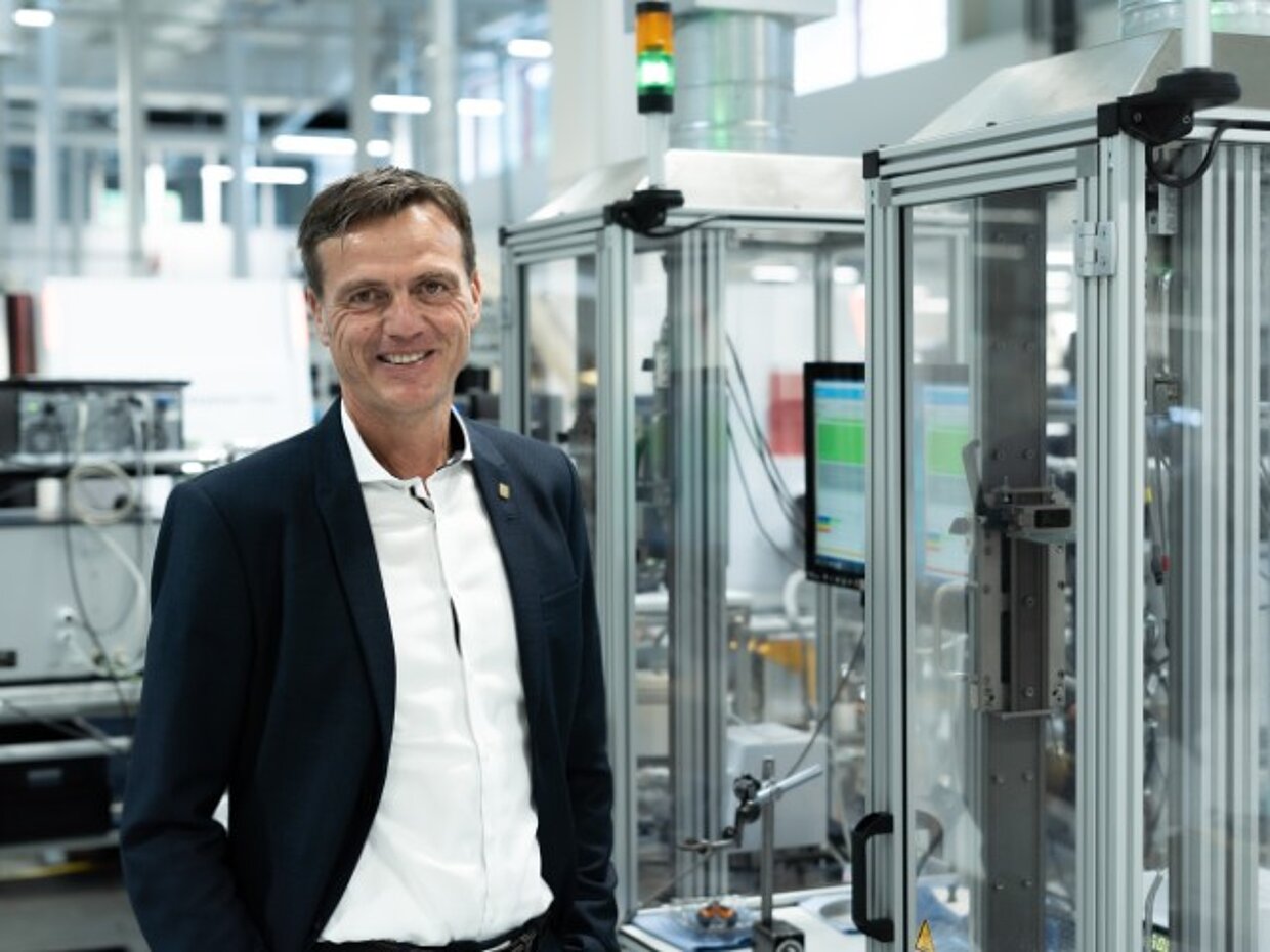 Ein Geschäftsmann steht in einem modernen Industriebereich neben Maschinen. Er trägt einen schwarzen Anzug und lächelt, während er in die Kamera schaut. Im Hintergrund sind Geräte und Monitoren sichtbar, die auf technologische Aktivitäten hinweisen.