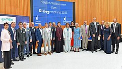 Eine Gruppe von 20 Personen steht lächelnd in einem modernen Raum. Im Hintergrund ist ein Banner mit der Aufschrift "Herzlich Willkommen zum Dialogempfang 2023" zu sehen. Alle tragen formelle Kleidung und scheinen an einem sozialen oder geschäftlichen Event teilzunehmen.